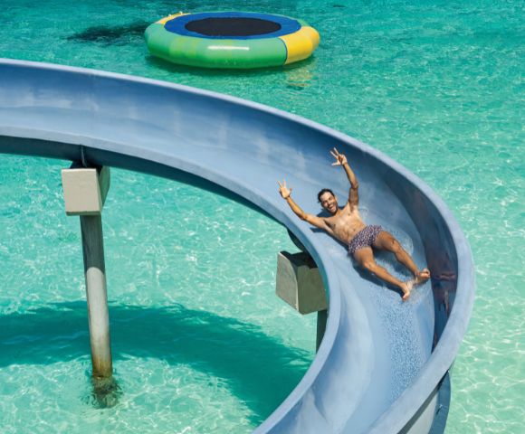 Man sliding down a blue waterslide into clear turquoise water, smiling and raising two fingers in a peace sign. Nearby, a colorful floating trampoline.