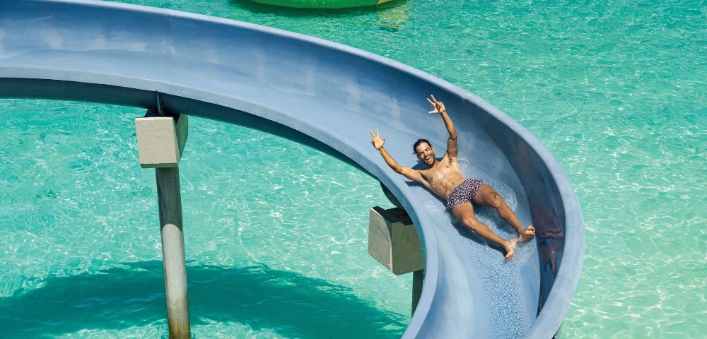 Man sliding down a blue waterslide into clear turquoise water, smiling and raising two fingers in a peace sign. Nearby, a colorful floating trampoline.