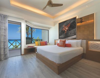 A serene hotel room features a large bed with colorful pillows, modern art on the wall, and a ceiling fan. A door opens to a balcony with a sea view.