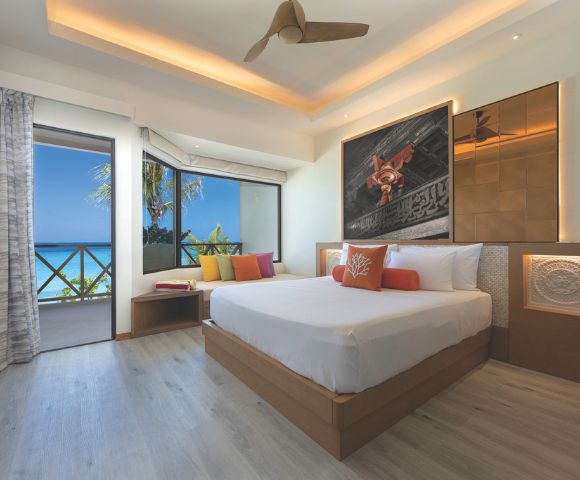 A serene hotel room features a large bed with colorful pillows, modern art on the wall, and a ceiling fan. A door opens to a balcony with a sea view.