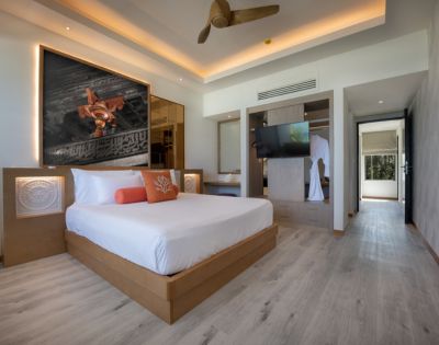 Modern hotel room with a king-sized bed, art above headboard, and warm lighting. Features a ceiling fan, wall-mounted TV, and light wood flooring.