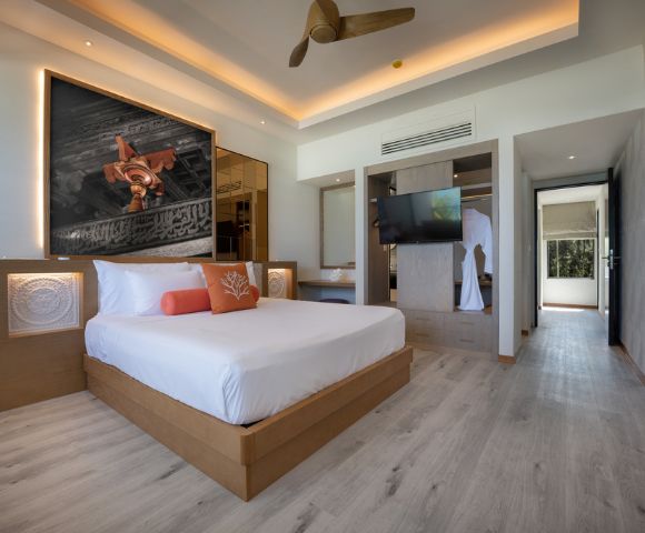 Modern hotel room with a king-sized bed, art above headboard, and warm lighting. Features a ceiling fan, wall-mounted TV, and light wood flooring.