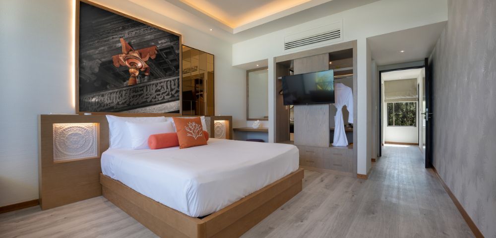 Modern hotel room with a king-sized bed, art above headboard, and warm lighting. Features a ceiling fan, wall-mounted TV, and light wood flooring.