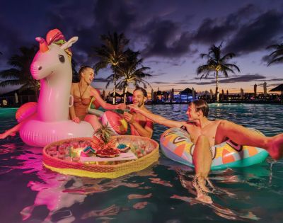 Three people relax on colorful inflatables in a pool at sunset, with palm trees silhouetted behind. They share drinks from a floating tray, creating a joyful vibe.