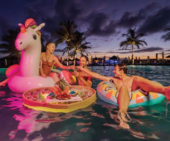 Three people relax on colorful inflatables in a pool at sunset, with palm trees silhouetted behind. They share drinks from a floating tray, creating a joyful vibe.