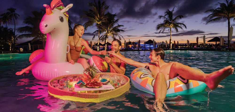 Three people relax on colorful inflatables in a pool at sunset, with palm trees silhouetted behind. They share drinks from a floating tray, creating a joyful vibe.