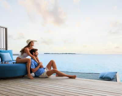 A couple relaxes on a deck overlooking a serene ocean, with light clouds in the sky. The scene conveys peace and contentment, with a cozy, intimate vibe.