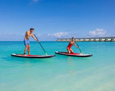 A man and woman enjoy paddleboarding on clear blue ocean water under a bright sky. Overwater bungalows are visible in the background, conveying a tropical vibe.