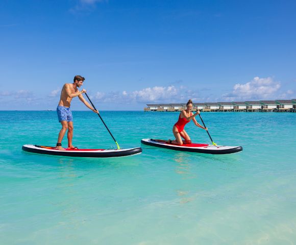 A man and woman enjoy paddleboarding on clear blue ocean water under a bright sky. Overwater bungalows are visible in the background, conveying a tropical vibe.