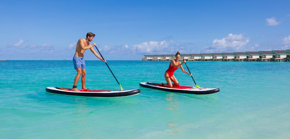 A man and woman enjoy paddleboarding on clear blue ocean water under a bright sky. Overwater bungalows are visible in the background, conveying a tropical vibe.