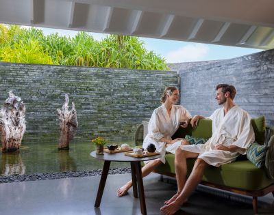A couple in white robes sits on a green couch in a spa-like setting, facing each other and smiling. They're beside a tranquil water feature with lush greenery and stone wall.