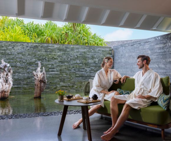 A couple in white robes sits on a green couch in a spa-like setting, facing each other and smiling. They're beside a tranquil water feature with lush greenery and stone wall.