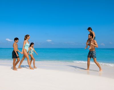 A family of five enjoys a sunny day on a pristine beach. Two adults and three children walk barefoot by the turquoise sea, appearing joyful and relaxed.