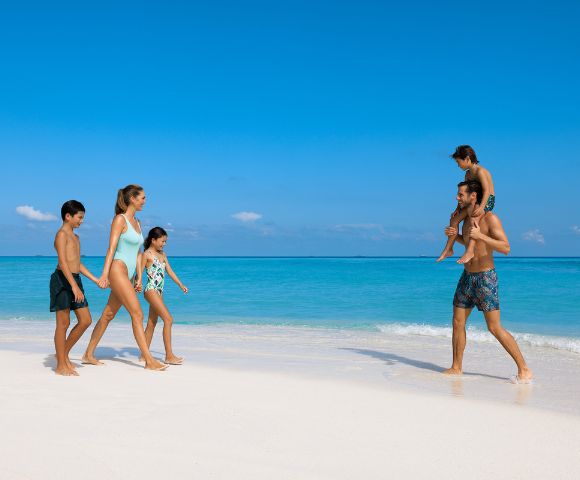 A family of five enjoys a sunny day on a pristine beach. Two adults and three children walk barefoot by the turquoise sea, appearing joyful and relaxed.