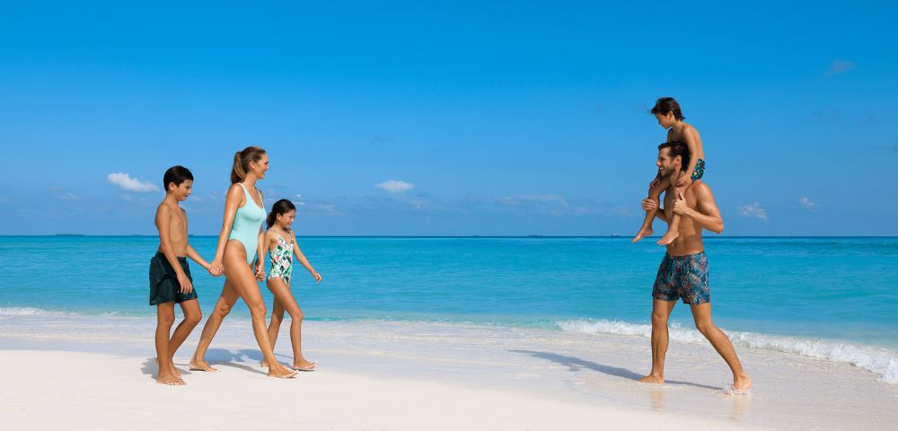 A family of five enjoys a sunny day on a pristine beach. Two adults and three children walk barefoot by the turquoise sea, appearing joyful and relaxed.
