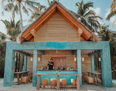 Tropical outdoor bar with a thatched roof, surrounded by palm trees. A bartender stands behind a turquoise counter, creating a relaxed, resort vibe.