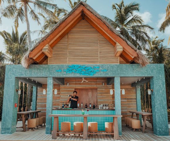 Tropical outdoor bar with a thatched roof, surrounded by palm trees. A bartender stands behind a turquoise counter, creating a relaxed, resort vibe.