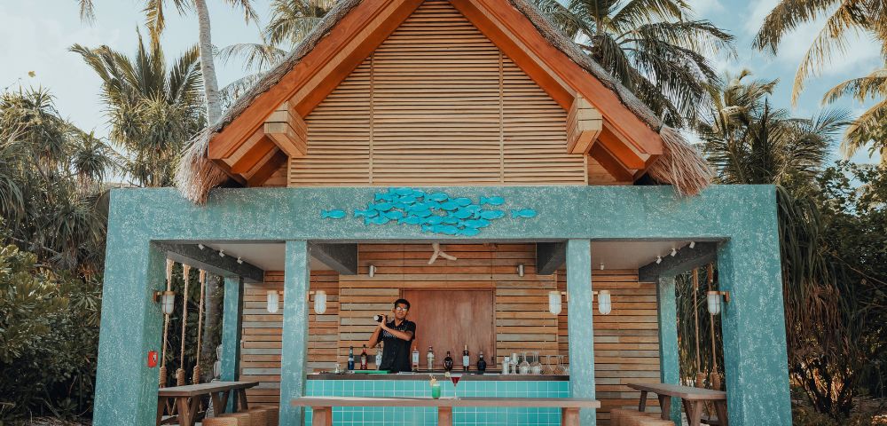 Tropical outdoor bar with a thatched roof, surrounded by palm trees. A bartender stands behind a turquoise counter, creating a relaxed, resort vibe.