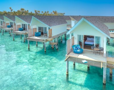 Overwater bungalows with wooden decks sit above clear turquoise waters. Each has lounge chairs, evoking a serene, tropical getaway. Sunny and tranquil setting.