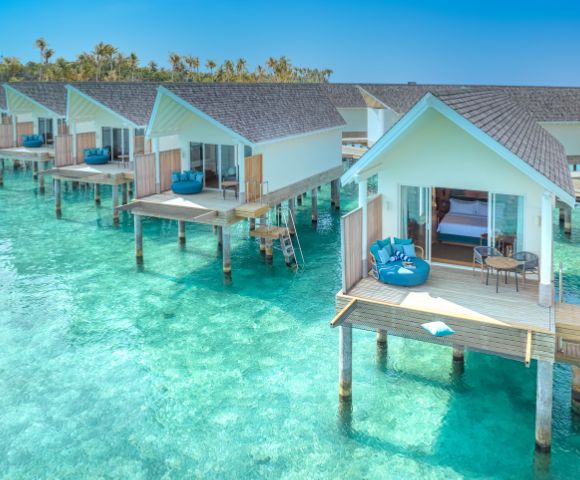 Overwater bungalows with wooden decks sit above clear turquoise waters. Each has lounge chairs, evoking a serene, tropical getaway. Sunny and tranquil setting.