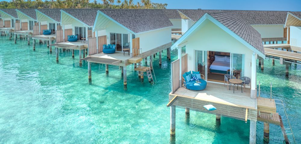 Overwater bungalows with wooden decks sit above clear turquoise waters. Each has lounge chairs, evoking a serene, tropical getaway. Sunny and tranquil setting.