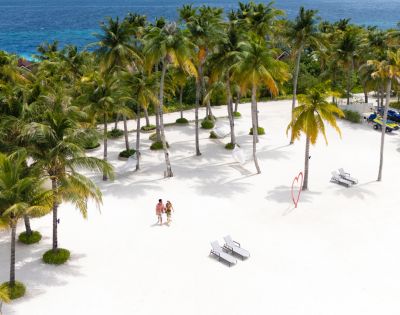 Aerial view of a sunny beach with white sand, palm trees, and blue ocean in the background. Two people walk near lounge chairs and a heart sculpture.