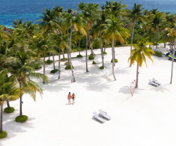 Aerial view of a sunny beach with white sand, palm trees, and blue ocean in the background. Two people walk near lounge chairs and a heart sculpture.