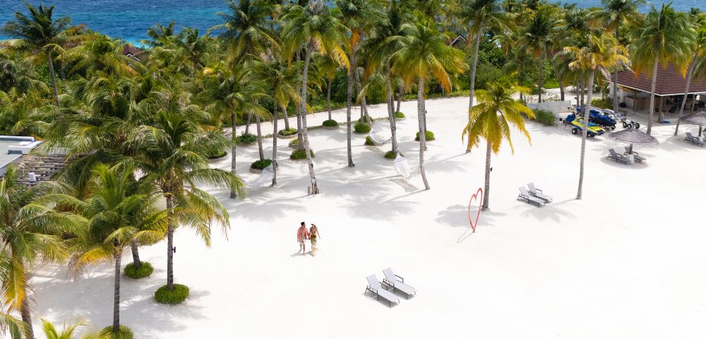 Aerial view of a sunny beach with white sand, palm trees, and blue ocean in the background. Two people walk near lounge chairs and a heart sculpture.