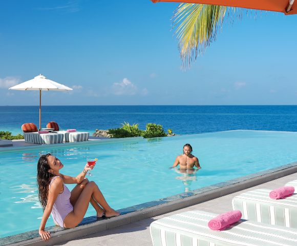 A woman in a swimsuit sits at an infinity pool's edge, holding a drink, smiling. A man swims nearby. Ocean view under a clear blue sky. Relaxed ambiance.