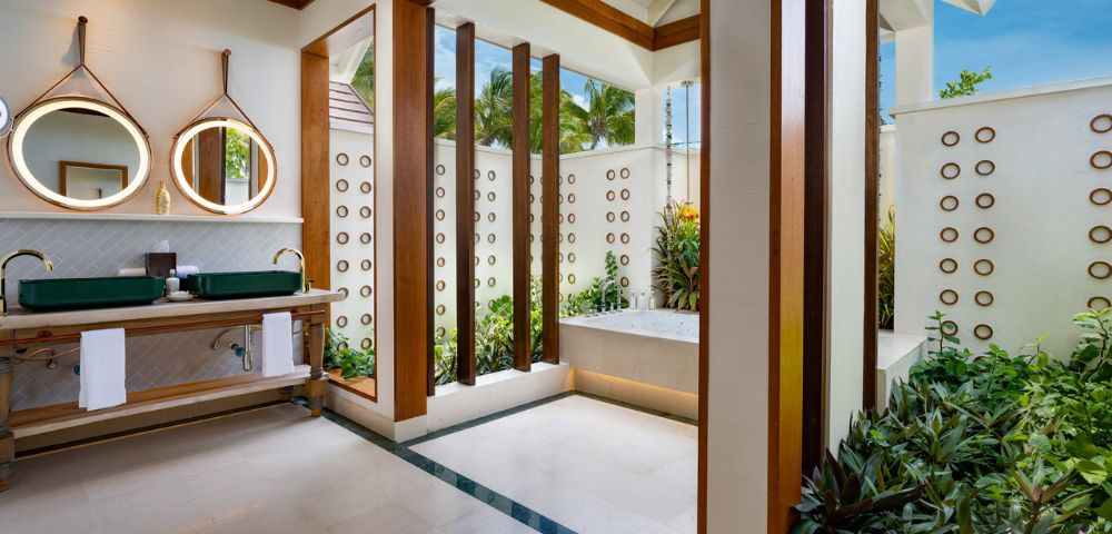 Bright, modern bathroom with open ceiling and lush plants. Features a tub, green countertop with dual sinks, and circular mirrors, creating a serene, spa-like ambiance.