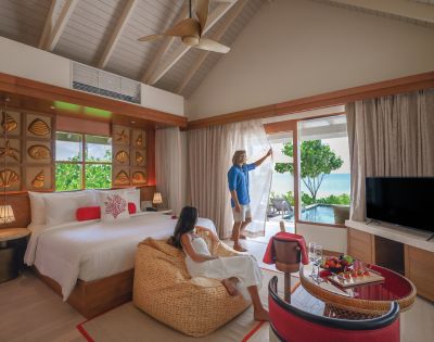 Cozy beachside room with a couple; a woman relaxes on a beanbag, while a man opens curtains to reveal a lush garden view and the ocean beyond.