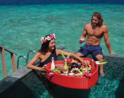 A woman with a floral crown and a man are in a pool, enjoying a floating breakfast in a red basket. The ocean is visible in the background, creating a serene setting.