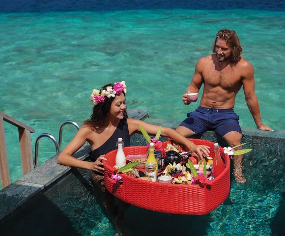 A woman with a floral crown and a man are in a pool, enjoying a floating breakfast in a red basket. The ocean is visible in the background, creating a serene setting.