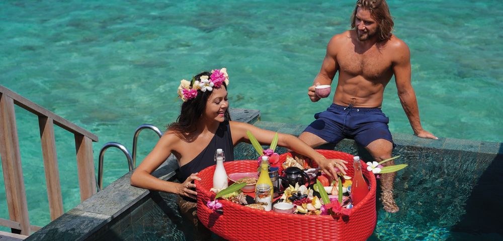 A woman with a floral crown and a man are in a pool, enjoying a floating breakfast in a red basket. The ocean is visible in the background, creating a serene setting.