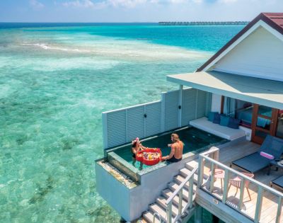 A couple enjoys a floating breakfast in a pool at an overwater villa, surrounded by clear turquoise sea, under a bright, sunny sky, evoking tranquility.