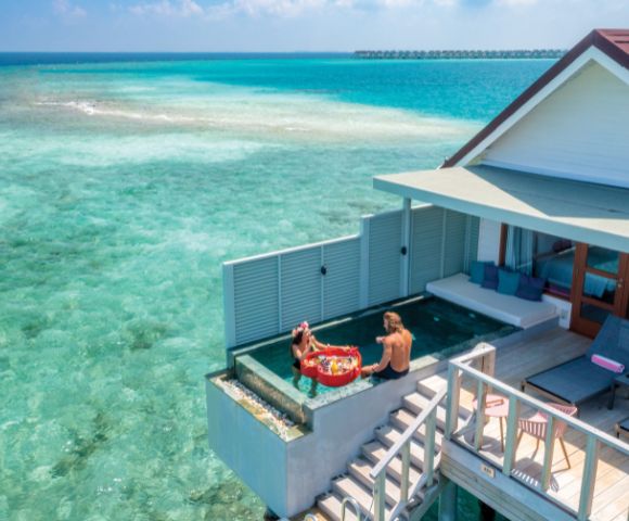 A couple enjoys a floating breakfast in a pool at an overwater villa, surrounded by clear turquoise sea, under a bright, sunny sky, evoking tranquility.
