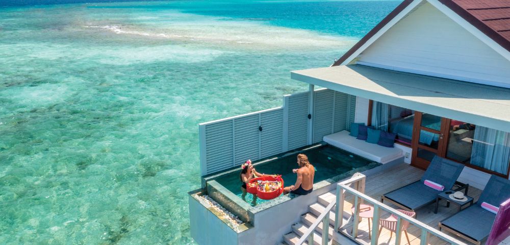 A couple enjoys a floating breakfast in a pool at an overwater villa, surrounded by clear turquoise sea, under a bright, sunny sky, evoking tranquility.