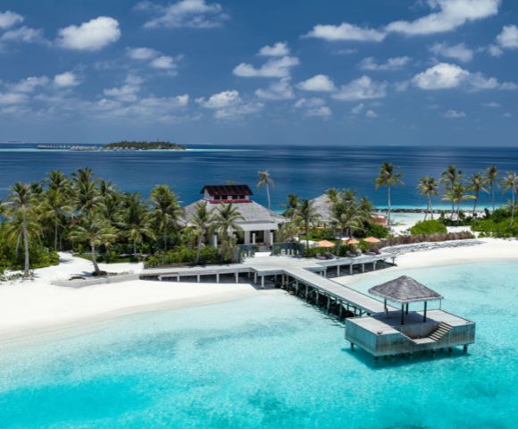 Tropical island resort with palm trees, white sand beaches, and turquoise waters. A long wooden pier leads to a gazebo, under a clear blue sky. Serene atmosphere.