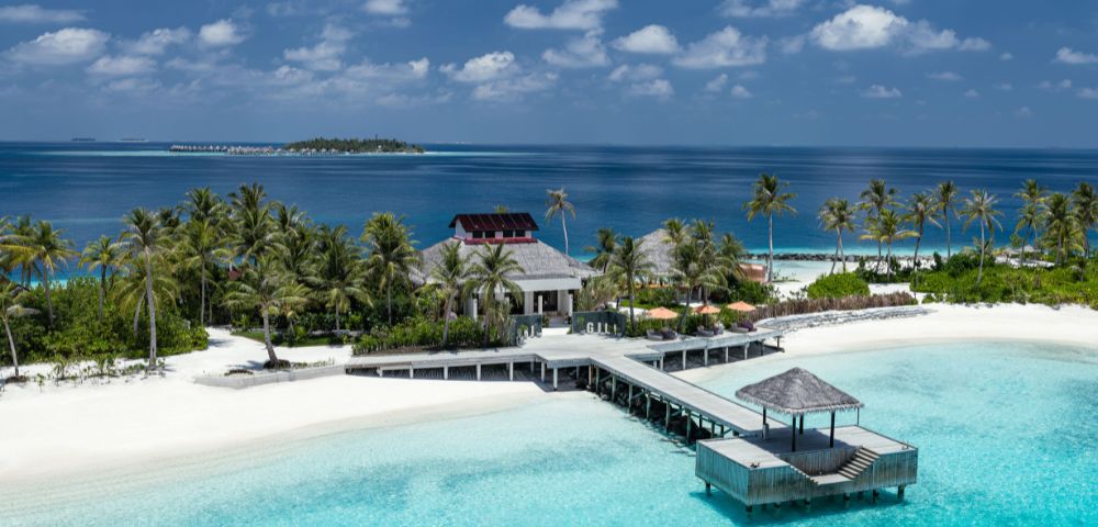 Tropical island resort with palm trees, white sand beaches, and turquoise waters. A long wooden pier leads to a gazebo, under a clear blue sky. Serene atmosphere.