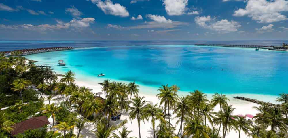 Tropical beach scene with turquoise ocean, white sand, and lush palm trees under a vibrant blue sky with scattered clouds. Peaceful and inviting atmosphere.
