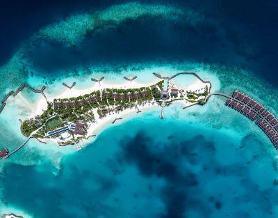 Aerial view of a tropical island resort surrounded by vibrant blue ocean. Overwater bungalows extend into the sea, with lush greenery and white sandy beaches.
