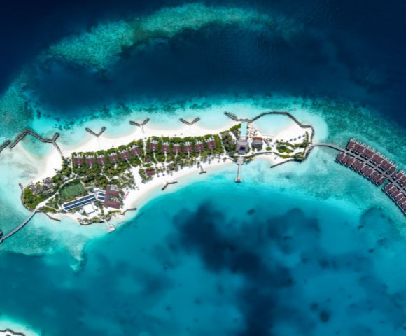 Aerial view of a tropical island resort surrounded by vibrant blue ocean. Overwater bungalows extend into the sea, with lush greenery and white sandy beaches.