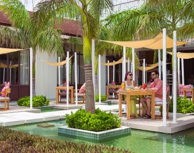 Two people dine outdoors at a tropical resort, seated at a table under palm trees and shade sails, next to a turquoise pool. Relaxed and sunny atmosphere.