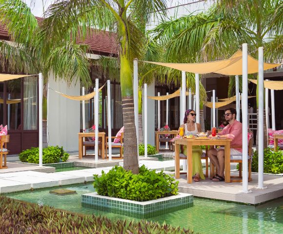 Two people dine outdoors at a tropical resort, seated at a table under palm trees and shade sails, next to a turquoise pool. Relaxed and sunny atmosphere.
