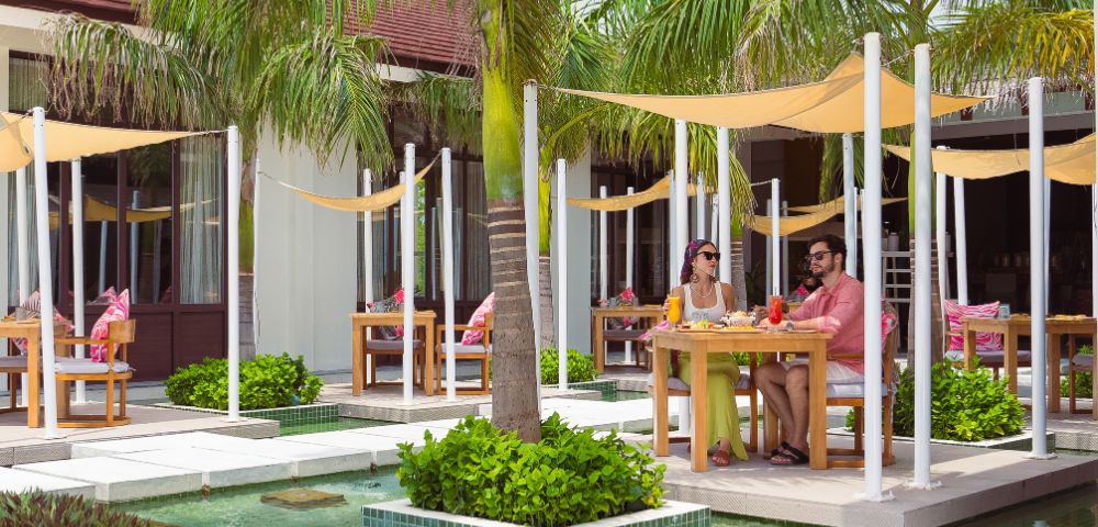 Two people dine outdoors at a tropical resort, seated at a table under palm trees and shade sails, next to a turquoise pool. Relaxed and sunny atmosphere.