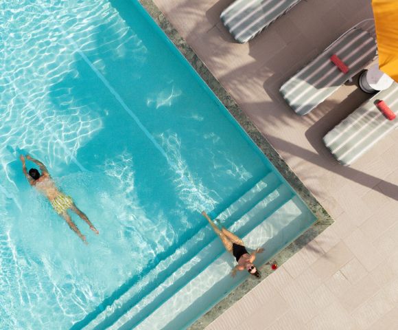 Two people enjoy a sunny day by a pool. One swims while the other relaxes on the steps. Nearby, lounge chairs and a yellow umbrella cast shadows.