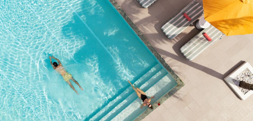 Two people enjoy a sunny day by a pool. One swims while the other relaxes on the steps. Nearby, lounge chairs and a yellow umbrella cast shadows.