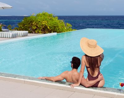 A couple relaxes at an infinity pool, overlooking the ocean. They're embracing; the woman wears a sun hat. The scene is tranquil and sunny, evoking a sense of leisure.