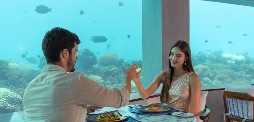 A couple toasts in an underwater restaurant with a view of fish swimming past large windows. The setting is romantic and serene.