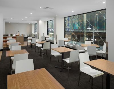 Bright, modern café with light wood tables and white chairs. Large windows reveal an urban street view, creating an open and inviting atmosphere.